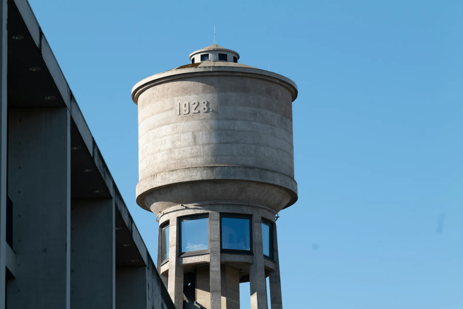 Torre de observação circular em concreto com anel superior e janelas, vista contra o céu azul, ao lado de uma estrutura metálica escura e detalhe com marcação “1923”.